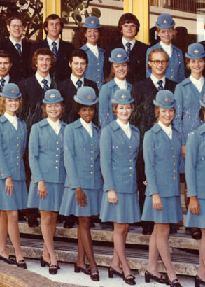 Flygv&auml;rdinnor och kabinpersonal i ljusbl&aring; uniformer och hattar. Personerna &auml;r uppst&auml;llda i flera rader framf&ouml;r en byggnad med glasd&ouml;rrar och guldramade f&ouml;nster.