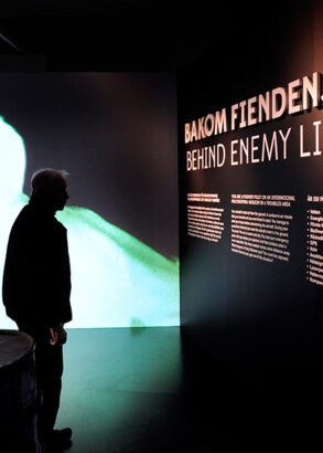 En bes&ouml;kare st&aring;r som en siluett i ett svagt upplyst rum framf&ouml;r informationstavlan "Bakom fiendens linje".