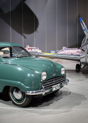 En turkos Saab 92 personbil parkerad bredvid ett mindre propellerplan i en utst&auml;llningshall.