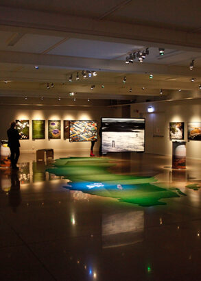 En gr&ouml;nbl&aring; matta i form av Sverige p&aring; golvet i en utst&auml;llningshall med tavlor och informationsplaketter p&aring; v&auml;ggarna. En bes&ouml;kare som besk&aring;dar utst&auml;llningen.