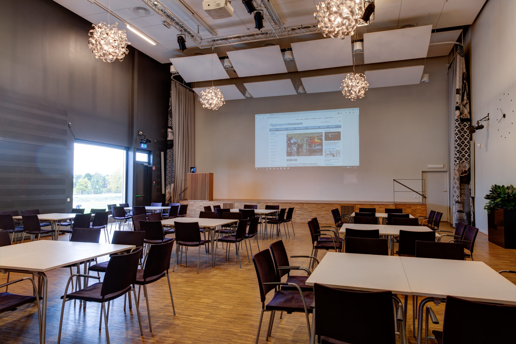 H&ouml;rsalen the saab wing har h&ouml;gt i tak med flera bord med stolar utplacerade nedanf&ouml;r en scen med sk&auml;rm.