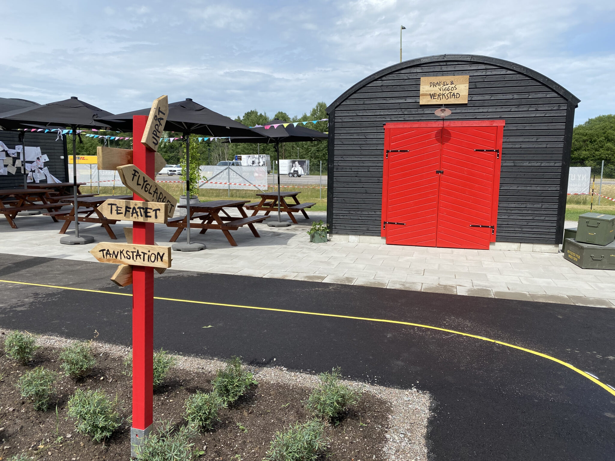 En v&auml;g f&ouml;r miniflygplan g&aring;r igenom lekplatsen Leklanda d&auml;r verkstaden med sin r&ouml;da dubbeld&ouml;rr &auml;r i fokus . Bredvid verkstaden st&aring;r flera bord med b&auml;nkar f&ouml;r den som vill &auml;ta sin mats&auml;ck utomhus.