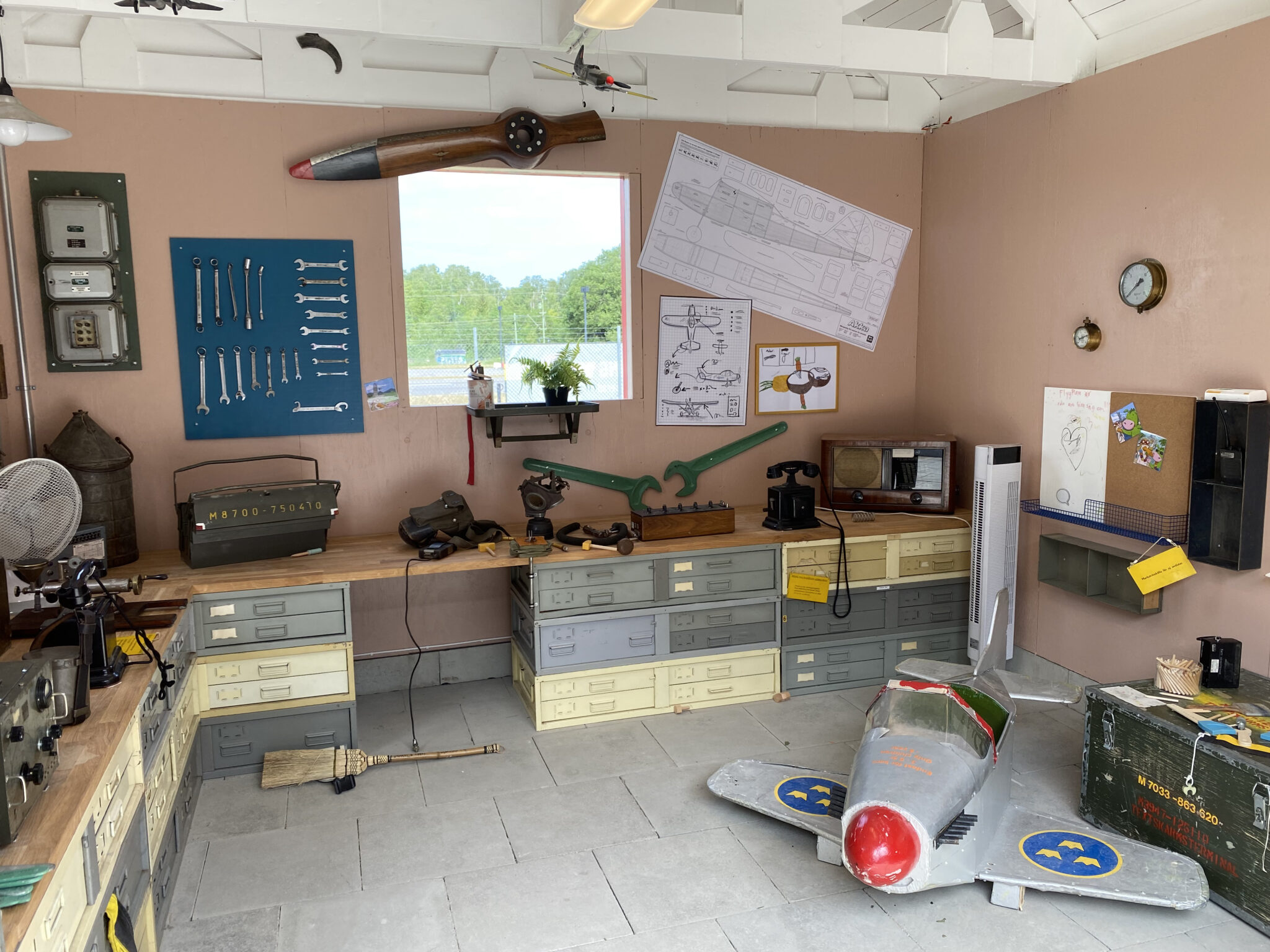 Inuti verkstaden st&aring;r ett flygplan och v&auml;ntar p&aring; reparation. P&aring; b&auml;nkarna och l&auml;ngs v&auml;ggarna finns verktyg och andra bra att ha saker n&auml;r man mekar med flygplan.