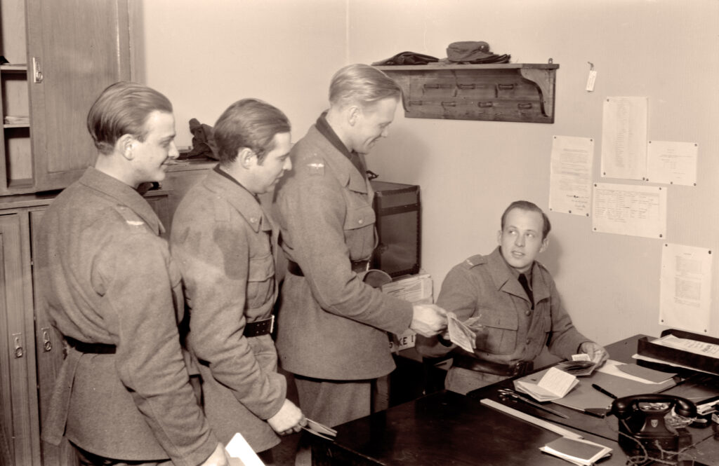 En svartvit arkivbild av tre uniformsklädda militärer på rad inne på ett kontor och en fjärde person vid ett skrivbord som betalar ut deras lön.