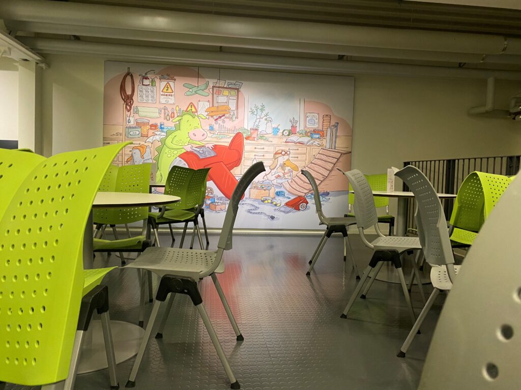 en hörna med bord och stolar där man kan sitta och äta sin medhavda matsäck i Flyglabbet. En stor tavla med maskotarna Drakel och Viggo i deras verkstad hänger på en av väggarna.