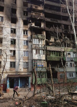 Tv&aring; &auml;ldre personer sopar utanf&ouml;r ing&aring;ngen till ett flerv&aring;ningshus. Flera v&aring;ningar &auml;r svarta efter brand och bara de nedre l&auml;genheterna verkar fortfarande ha f&ouml;nster kvar.