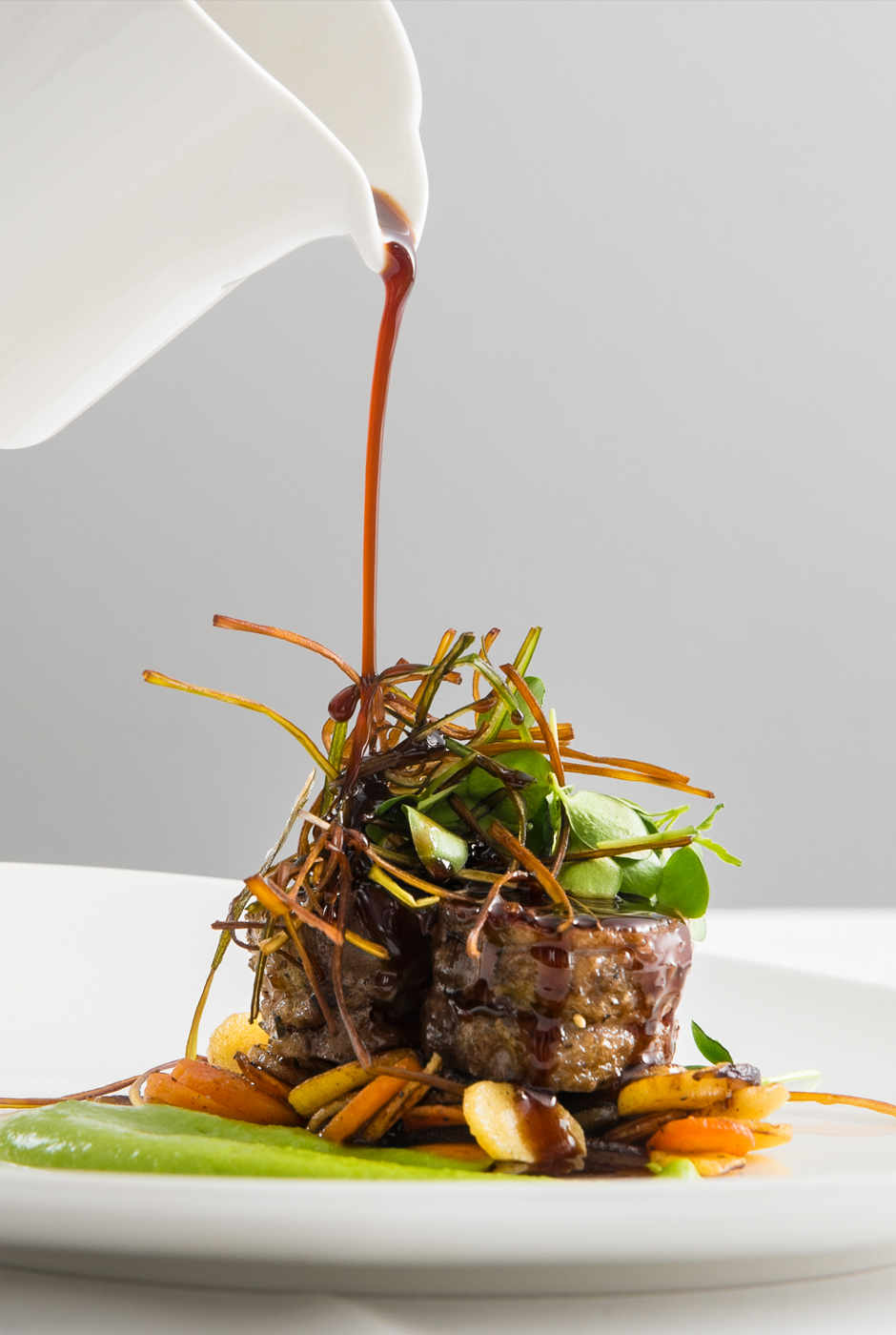 En k&ouml;ttbit vackert upplagd p&aring; en b&auml;dd av rotfrukter p&aring; en vit tallrik. En person h&auml;ller brun s&aring;s p&aring; toppen som &auml;r dekorerad med mer gr&ouml;nsaker.